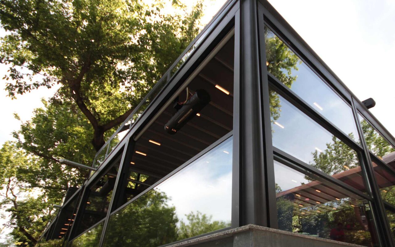 Otomatik giyotin cam sistemleri, dış mekanları dört mevsim kullanılabilir hale getiren motorlu cam sistemleridir. Bu görselde bir kafenin açık alanında uygulanan modern giyotin cam sistemi yer almaktadır.